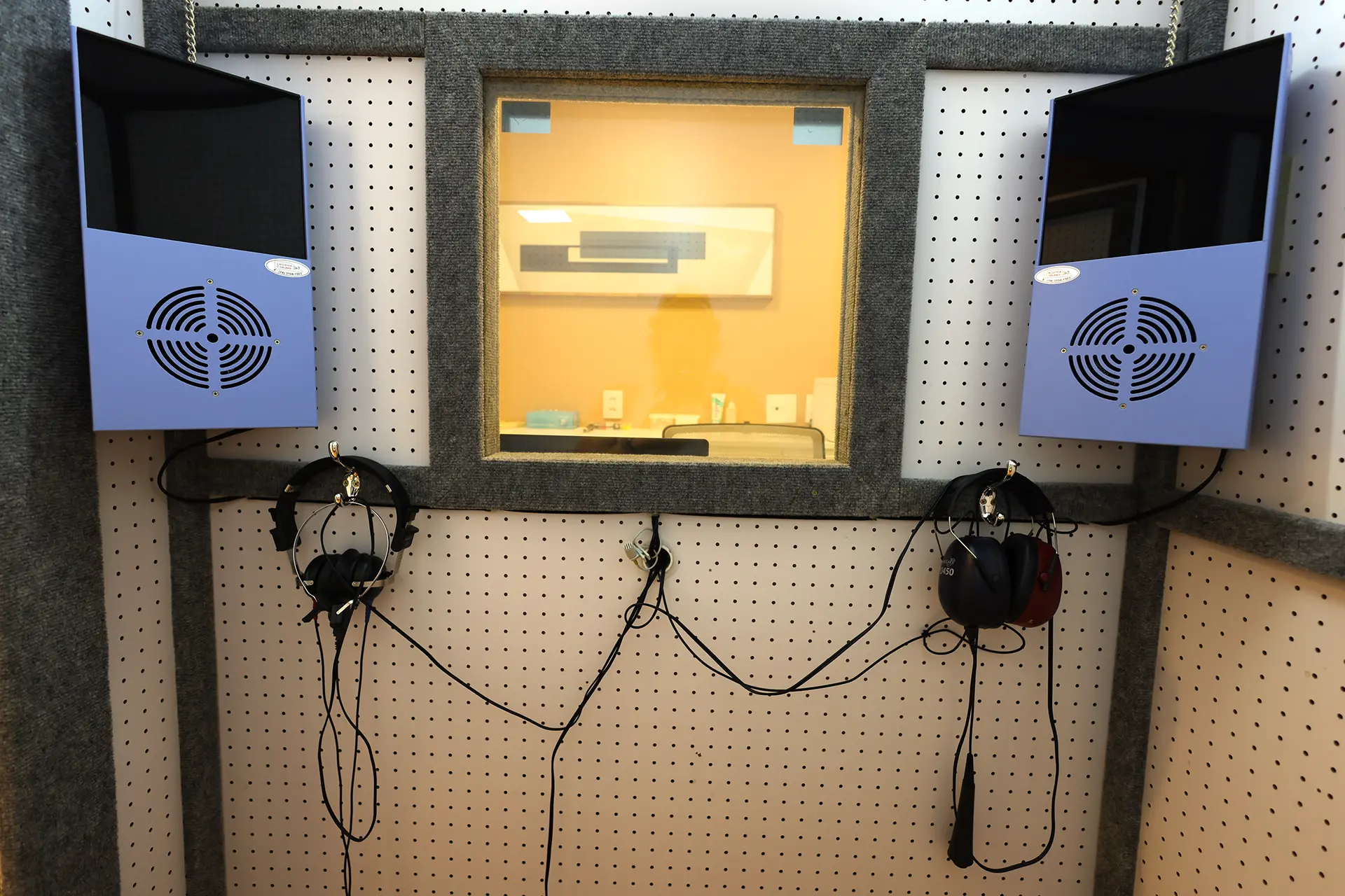 Interior da cabina acústica da Clínica OtoCare, em Brasília, com fones de audiometria, caixas acústicas e janela para a sala do fonoaudiólogo