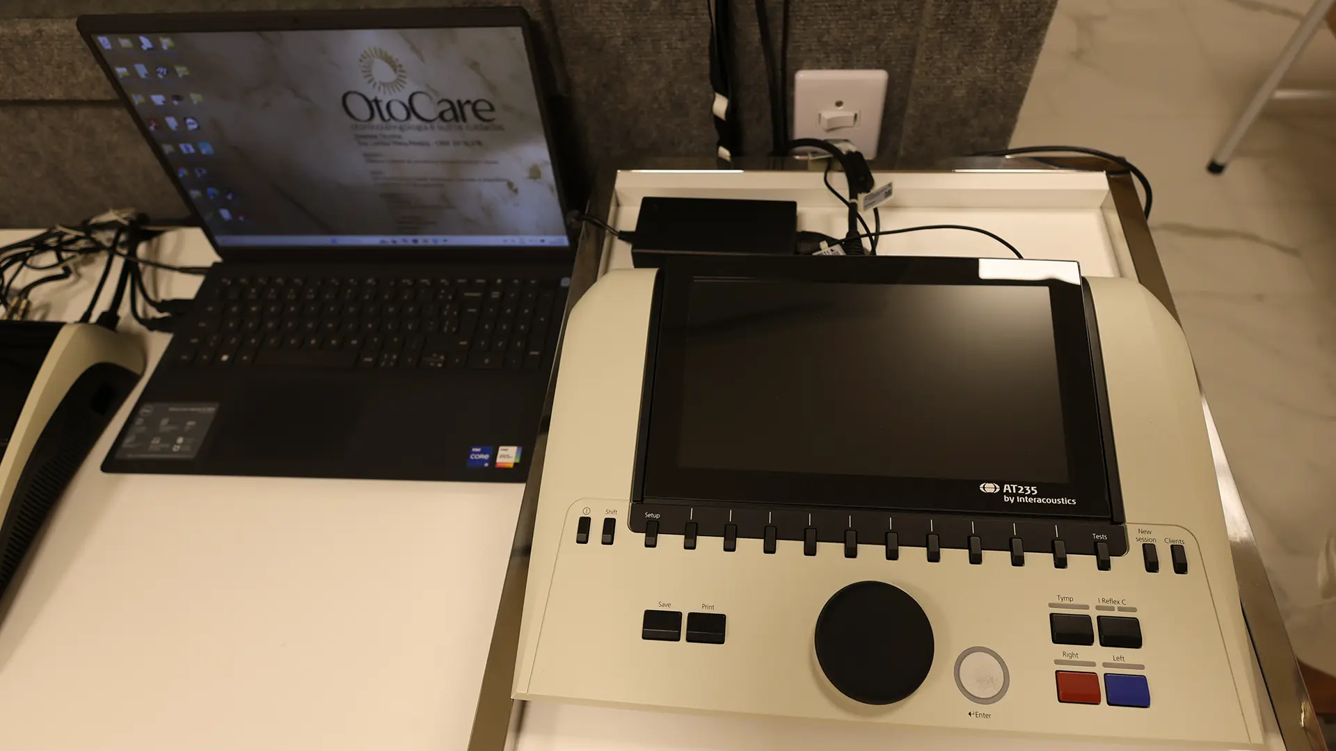 Impedanciômetro AT235 na sala de exames da Clínica OtoCare, com um notebook ao lado mostrando o logotipo da clínica