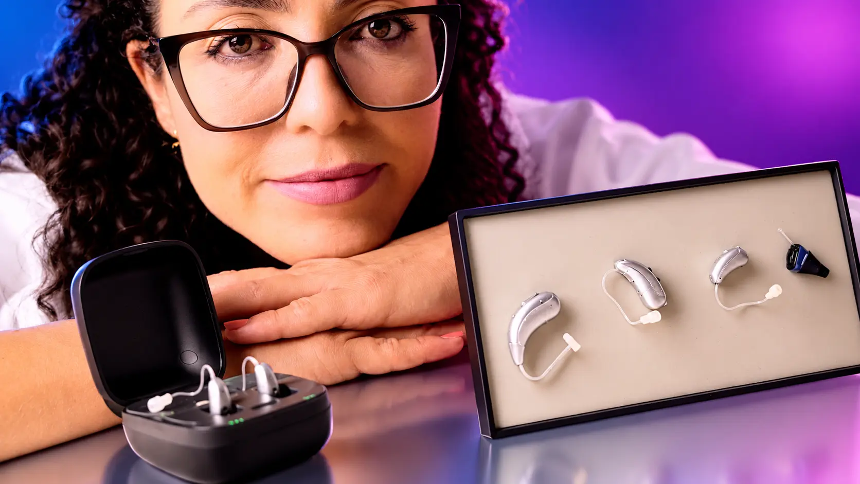 Dra. Larissa Vilela ao lado de quatro modelos de aparelho auditivo, retroauriculares e intracanal, e de um estojo carregador com um par de aparelhos
