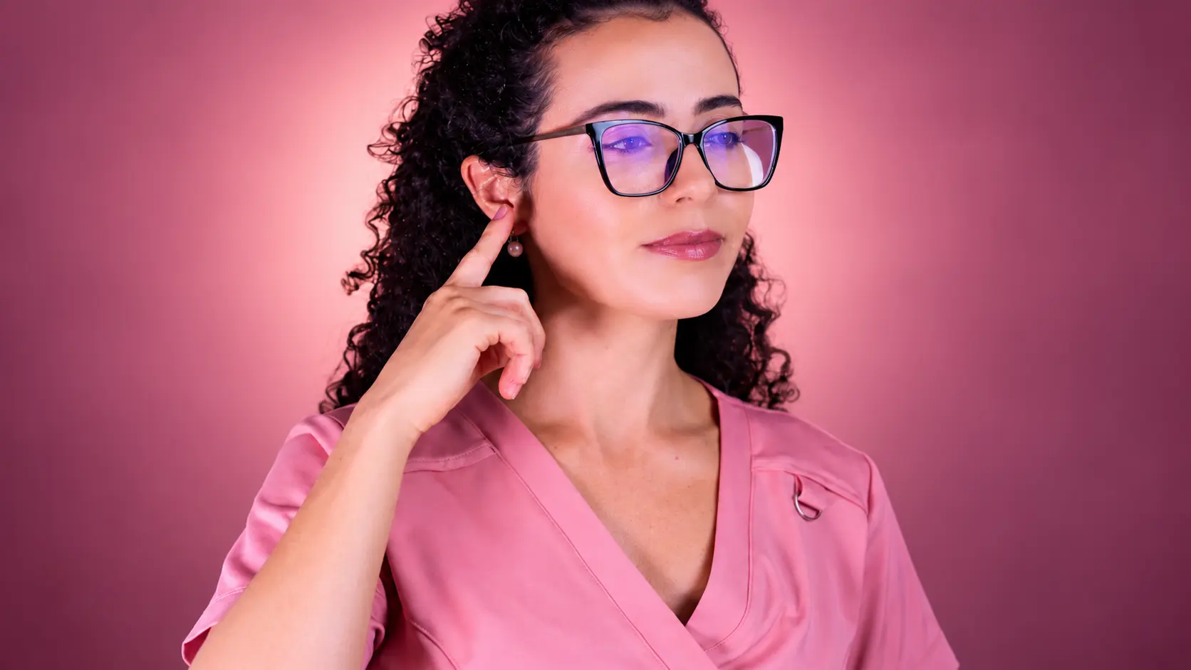 Mulher de óculos e jaleco rosa com o dedo apontando para a própria orelha, ilustrando a avaliação do grau de surdez para aposentadoria